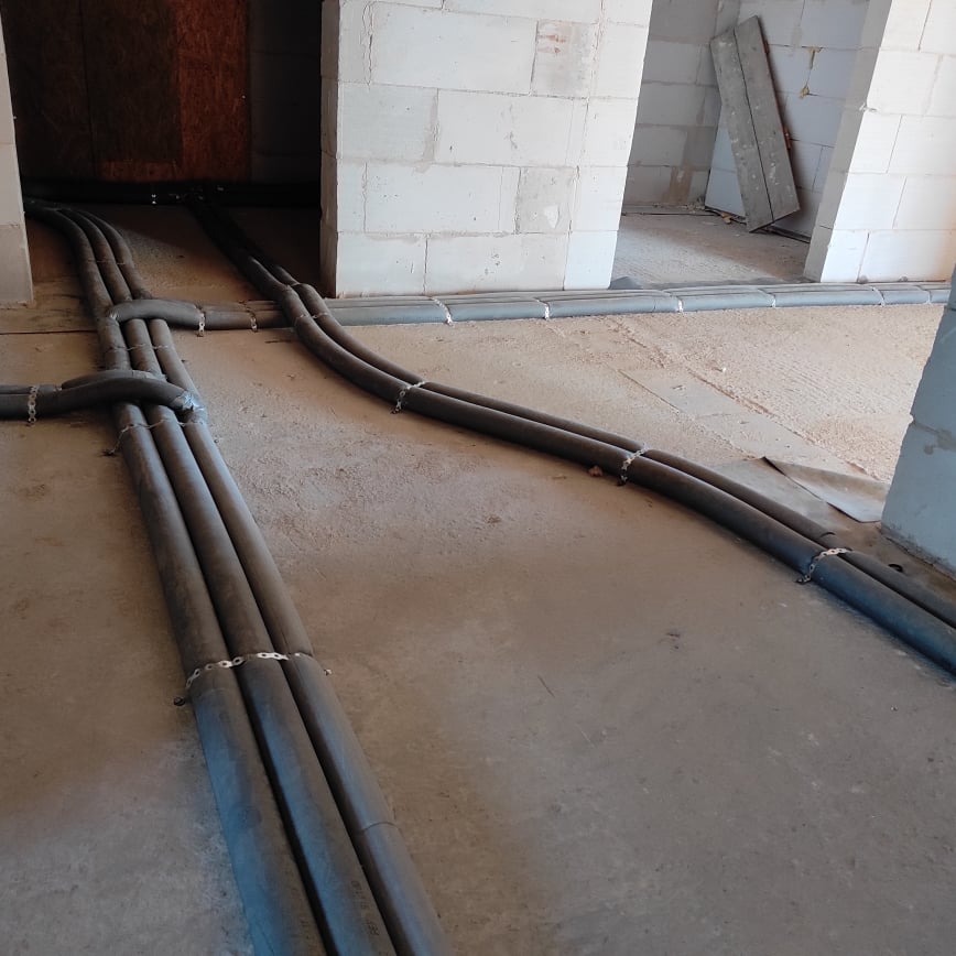 Szare rury instalacyjne prowadzone po betonowej podłodze w surowym wnętrzu budynku, mocowane metalowymi obejmami, widoczne ślady budowy i białe bloczki.