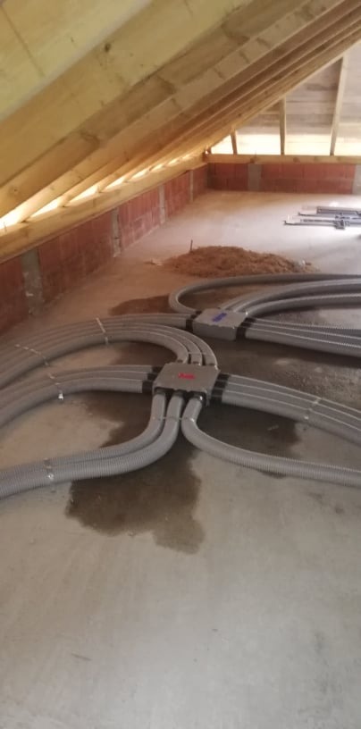 Szare, karbowane rury wentylacyjne rozchodzą się promieniście z centralnych, szarych skrzynek rozdzielczych na poddaszu w surowym stanie, widoczne drewniane krokwie dachu i ceglana ścianka kolankowa.