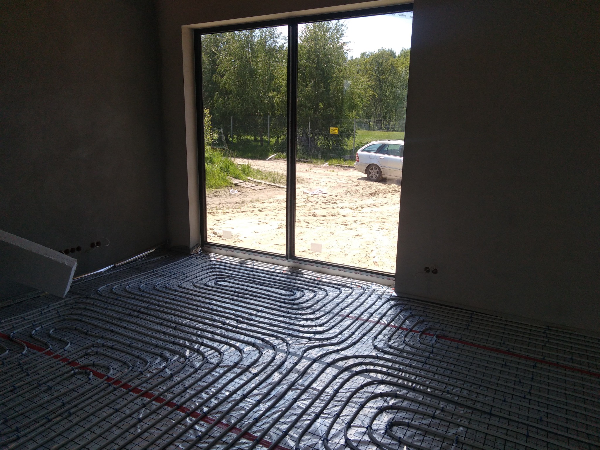 Instalacja ogrzewania podłogowego w trakcie realizacji w Łodzi, widoczne rury na folii izolacyjnej przed wylewką, z widokiem na podwórko przez duże okno.