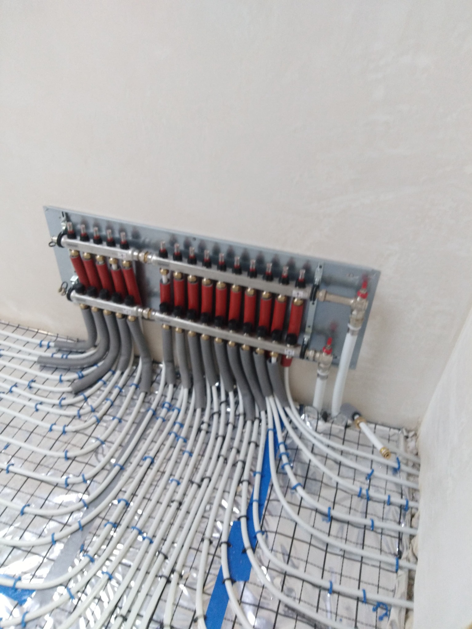 Rozdzielacz ogrzewania podłogowego z rurami ułożonymi na folii aluminiowej z siatką i przymocowanymi niebieskimi uchwytami, widok z góry.