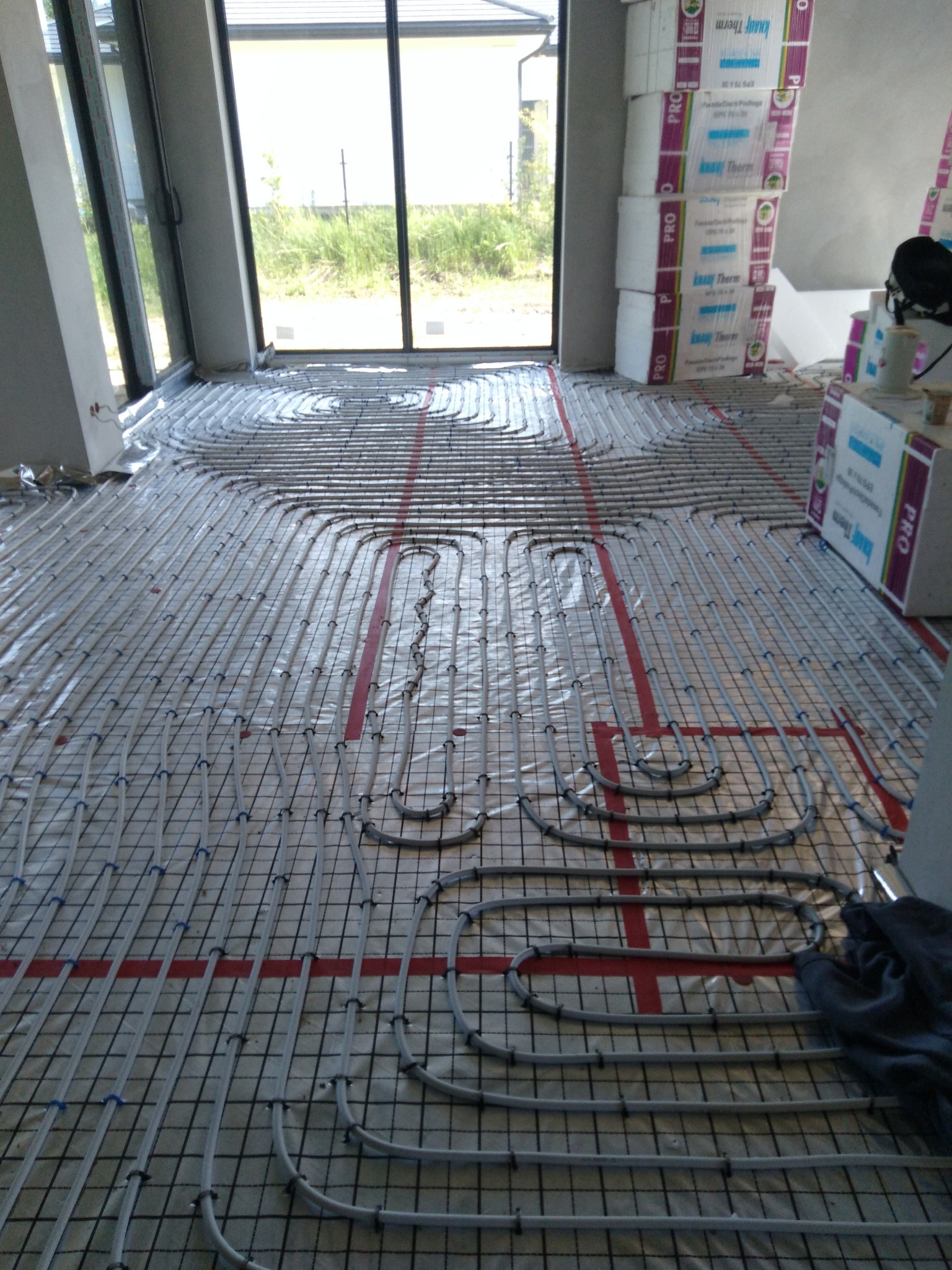 Szare rury ogrzewania podłogowego ułożone na folii z siatką, widoczne czerwone taśmy i styropianowe płyty izolacyjne w surowym wnętrzu domu w trakcie budowy.