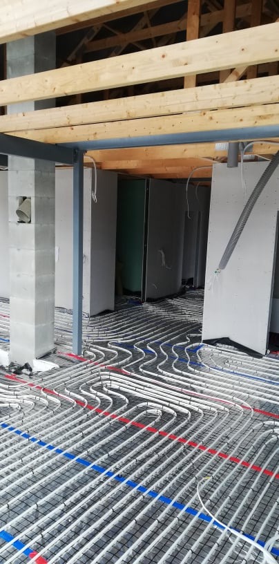 Białe rurki ogrzewania podłogowego ułożone na czarnej siatce, widoczne elementy konstrukcyjne budynku: drewniane belki stropowe i betonowe słupy.