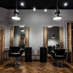Wnętrze salonu fryzjerskiego w stylu industrialnym z dwoma stanowiskami: lustra oprawione w surowe drewno, czarne fotele fryzjerskie, lampy wiszące z metalowymi kloszami i parkiet w jodełkę.