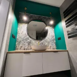 Nowoczesna łazienka z okrągłym lustrem, umywalką nablatową i zieloną ścianą z motywem roślinnym, oświetlona złotymi lampami wiszącymi.