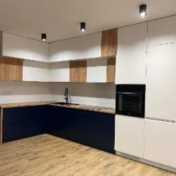 Nowoczesna kuchnia z granatowymi szafkami dolnymi, drewnianymi blatami, białymi szafkami górnymi z drewnianymi wstawkami i białymi płytkami na ścianie.