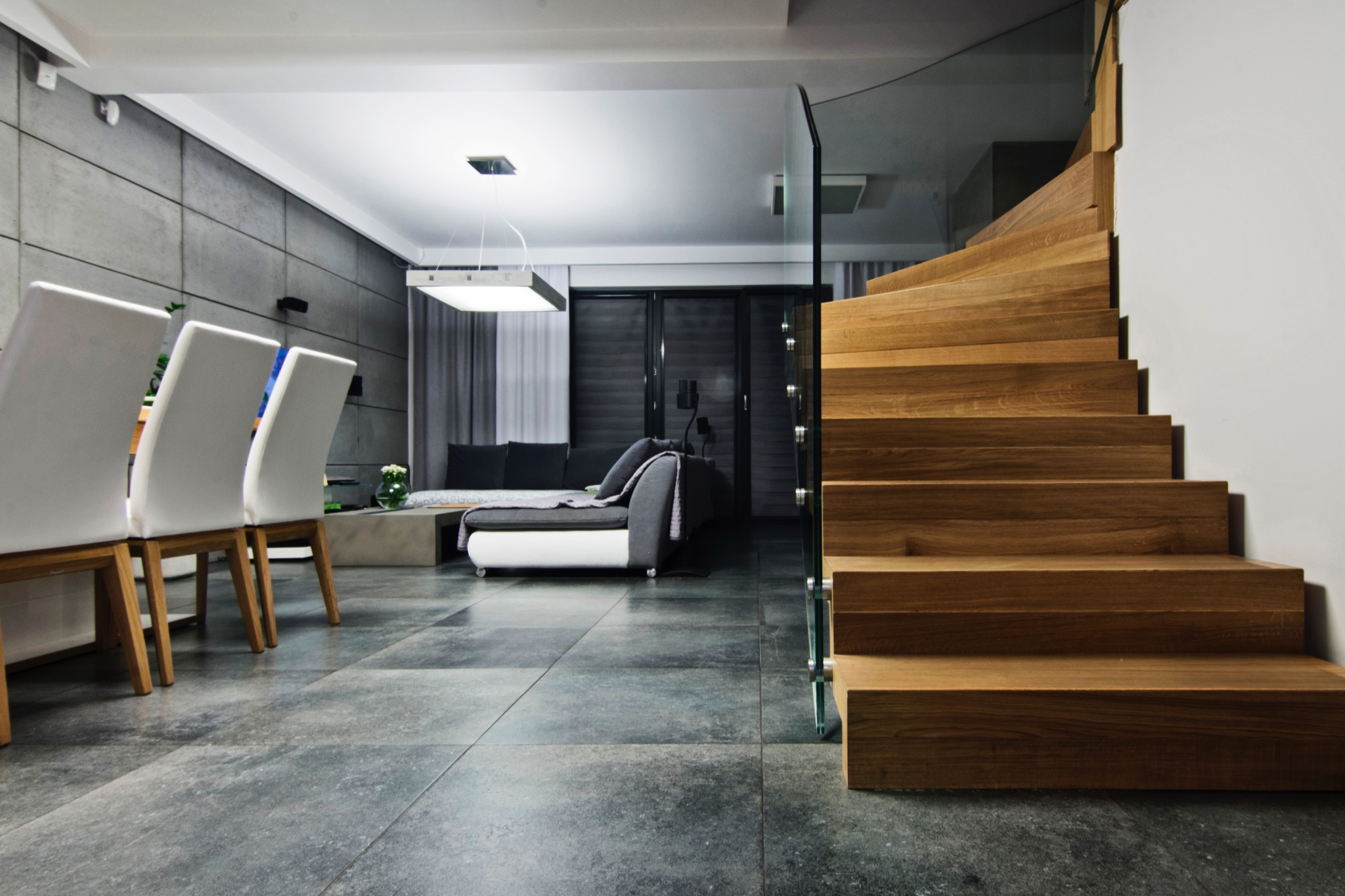 Nowoczesne wnętrze: szare płytki podłogowe, drewniane schody z szklaną balustradą, białe krzesła i sofa w tle, minimalistyczne oświetlenie.