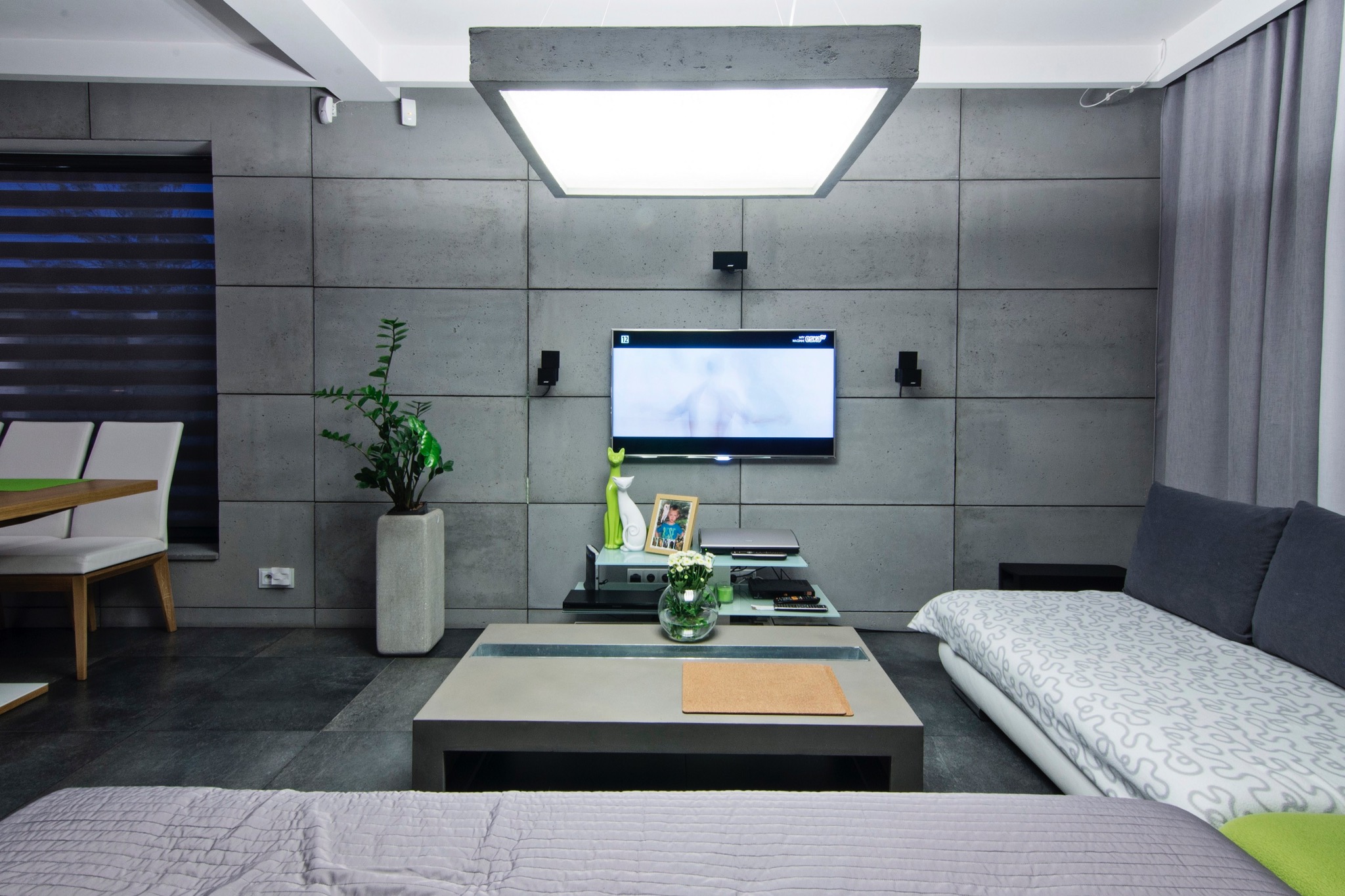 Nowoczesny salon z szarą betonową ścianą z paneli, kwadratową lampą sufitową, niskim stolikiem kawowym i sofą z wzorzystą tapicerką.