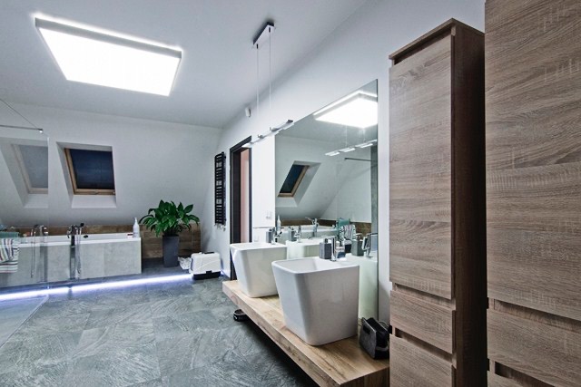 Jasna łazienka na poddaszu ze skośnym oknem, dwoma białymi umywalkami na drewnianym blacie, szarą podłogą i wysoką szafką z okleiną drewnopodobną.