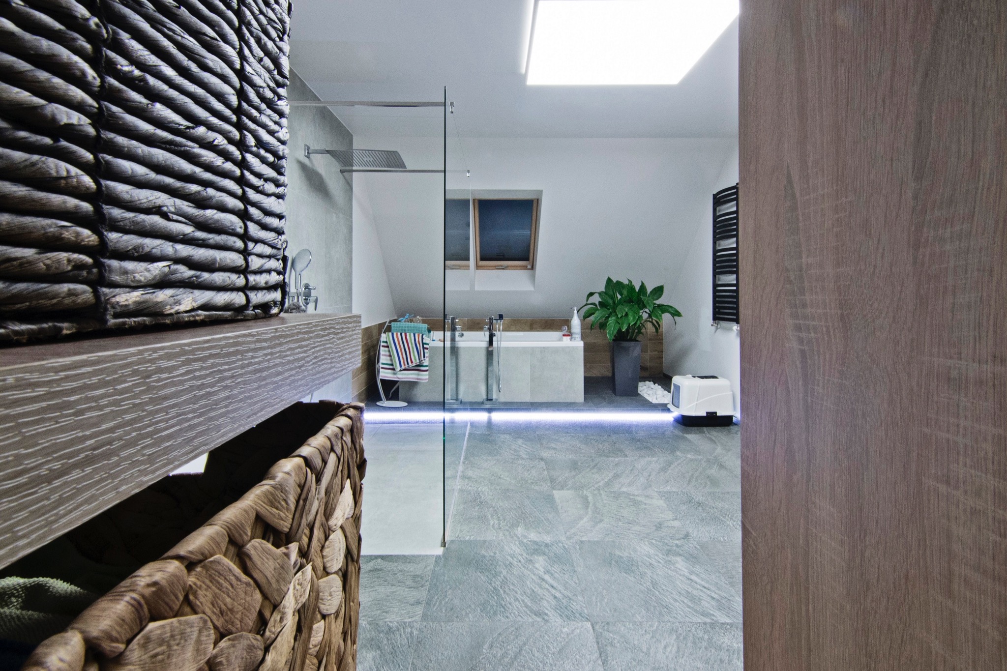 Nowoczesna łazienka na poddaszu z szarą płytką podłogową, szklaną kabiną prysznicową, oknem dachowym i dekoracyjnymi koszami z wikliny.