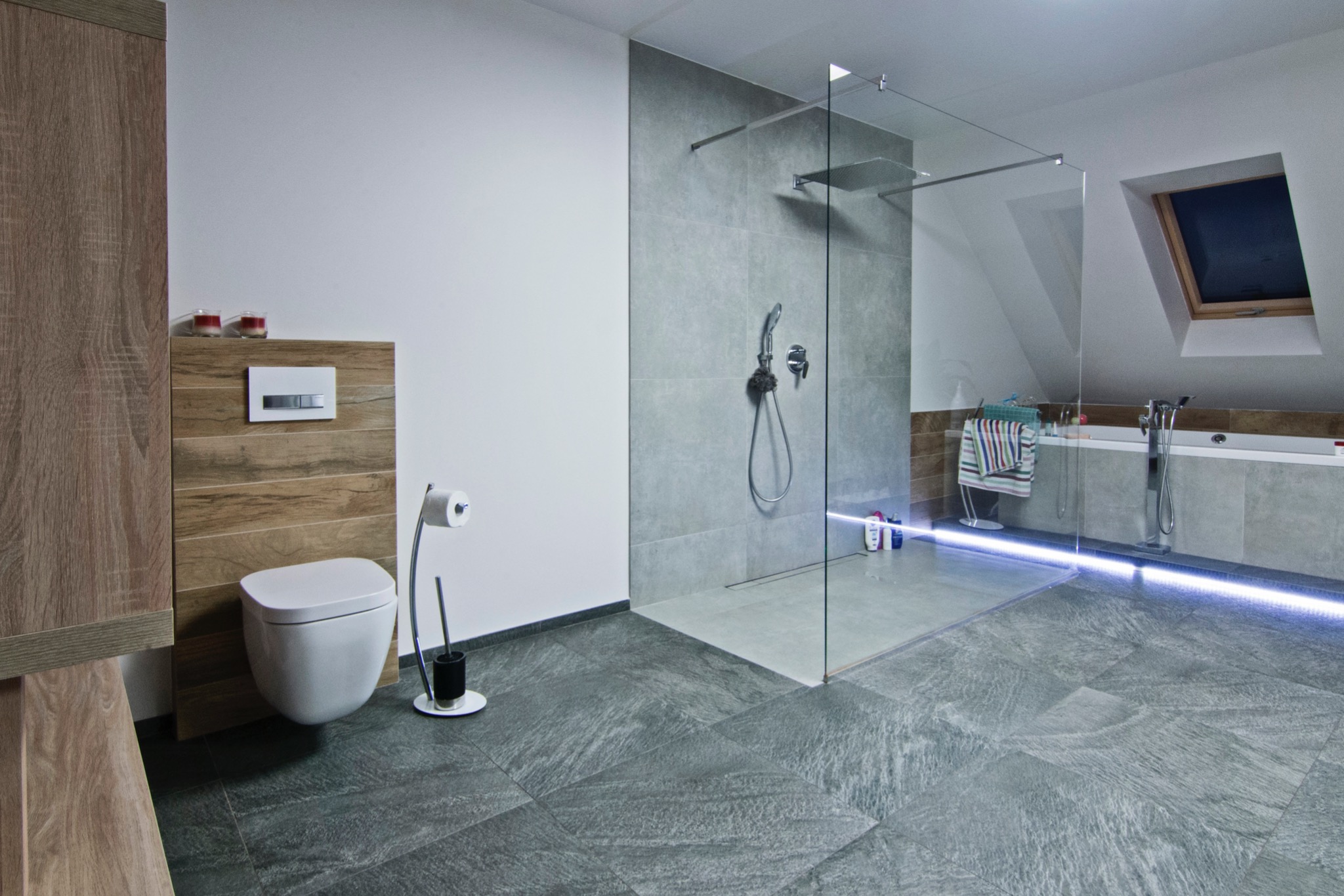 Nowoczesna łazienka na poddaszu z kabiną prysznicową walk-in, szarą płytką imitującą kamień na podłodze i ścianach, podświetleniem LED przy wannie oraz drewnianymi akcentami przy toalecie.