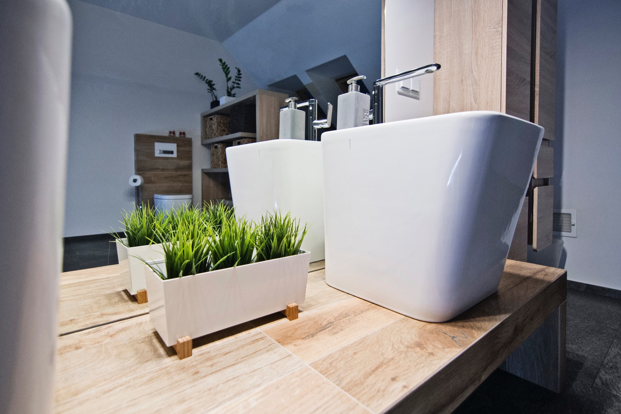 Nowoczesna łazienka z dwiema białymi umywalkami nablatowymi na drewnianym blacie, dekoracyjna trawa w doniczce, minimalistyczne dozowniki mydła, toaleta w tle.