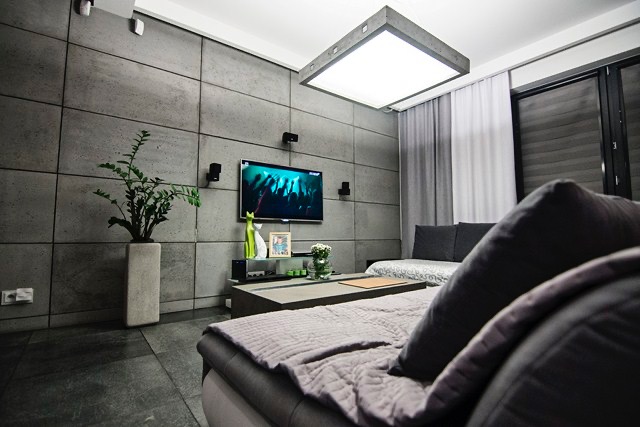 Nowoczesny salon z szarą ścianą z betonowych paneli, podświetlonym sufitem, telewizorem i minimalistycznymi dekoracjami, widok z perspektywy sofy z pikowaną narzutą.