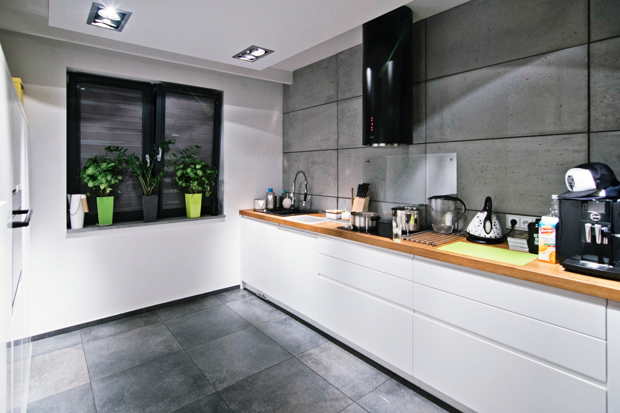 Nowoczesna kuchnia z białymi, minimalistycznymi szafkami, drewnianym blatem, szarą ścianą z betonowych płyt i ciemnoszarą podłogą z płytek. Na blacie czajnik, ekspres do kawy i naczynia. W oknie...