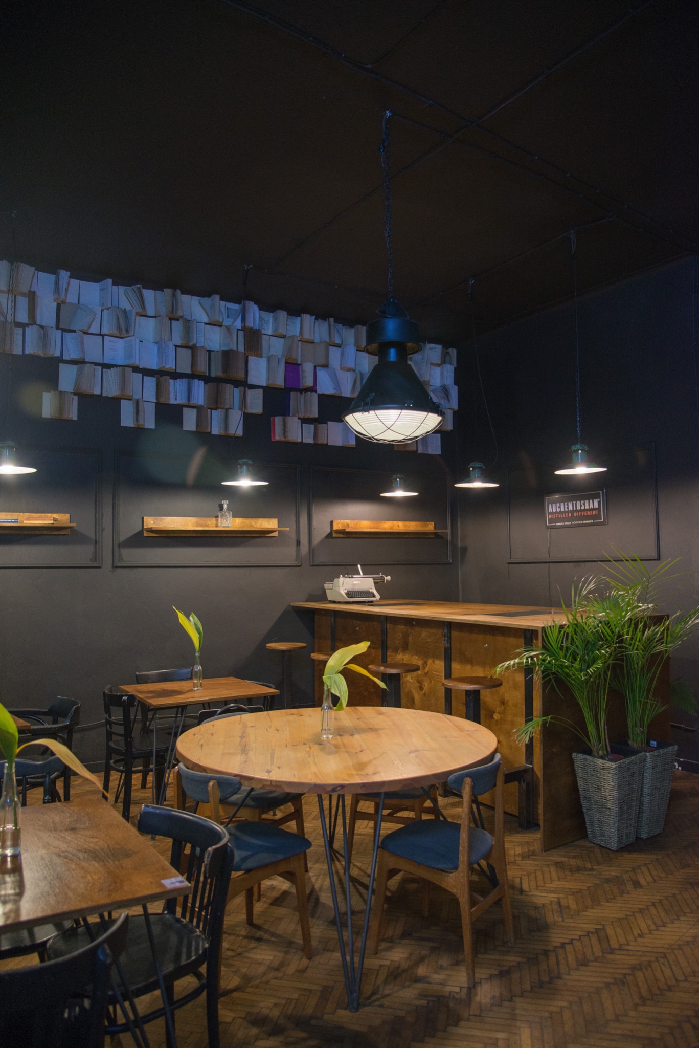 Wnętrze kawiarni w stylu retro z drewnianymi stolikami, krzesłami, barem, dekoracją z książek na ścianie i roślinami doniczkowymi.