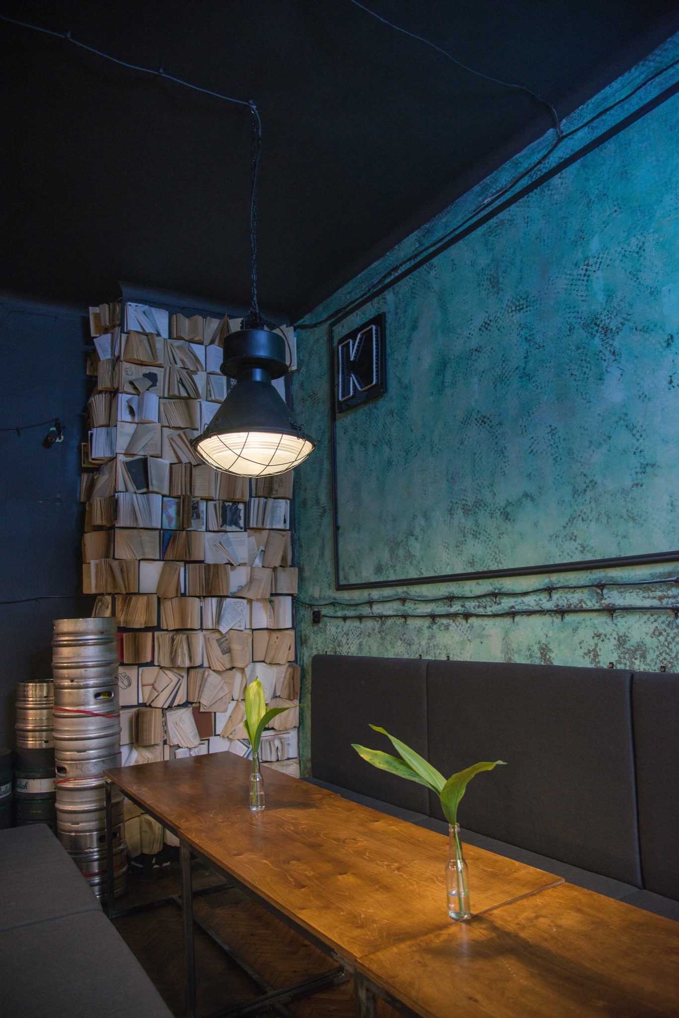 Industrialny wystrój baru: drewniany stół, ściana z książek, metalowe beczki, lampa wisząca i turkusowa ściana z neonem 'K'.