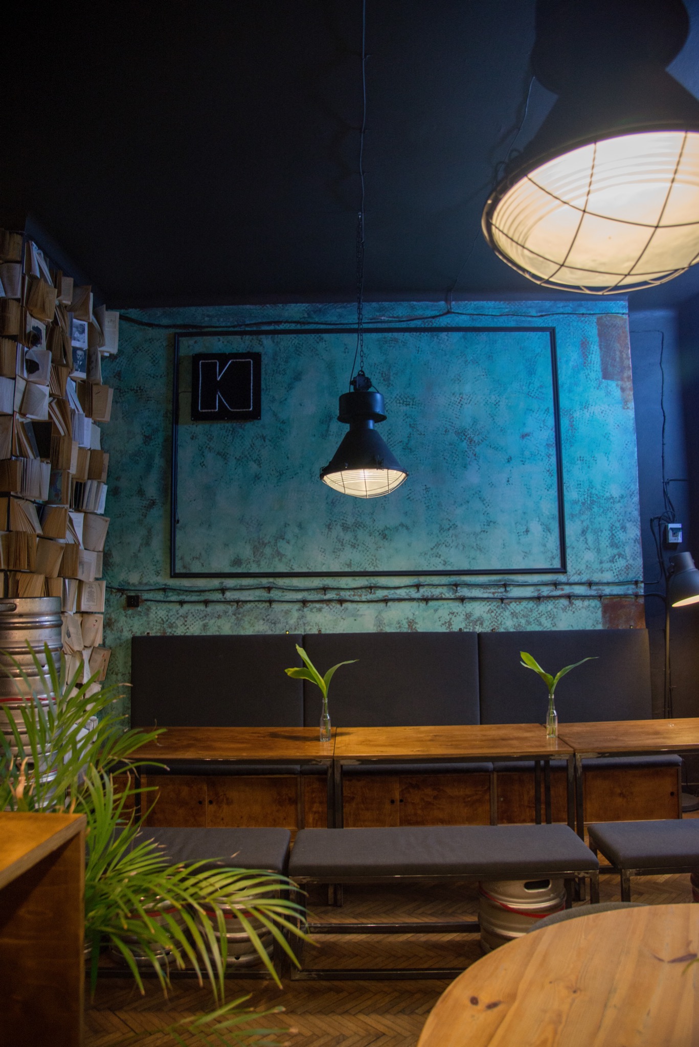 Industrialna aranżacja wnętrza kawiarni z turkusową ścianą, lampą wiszącą, drewnianymi stolikami i ławkami z szarymi poduszkami, dekoracją z książek i beczek.