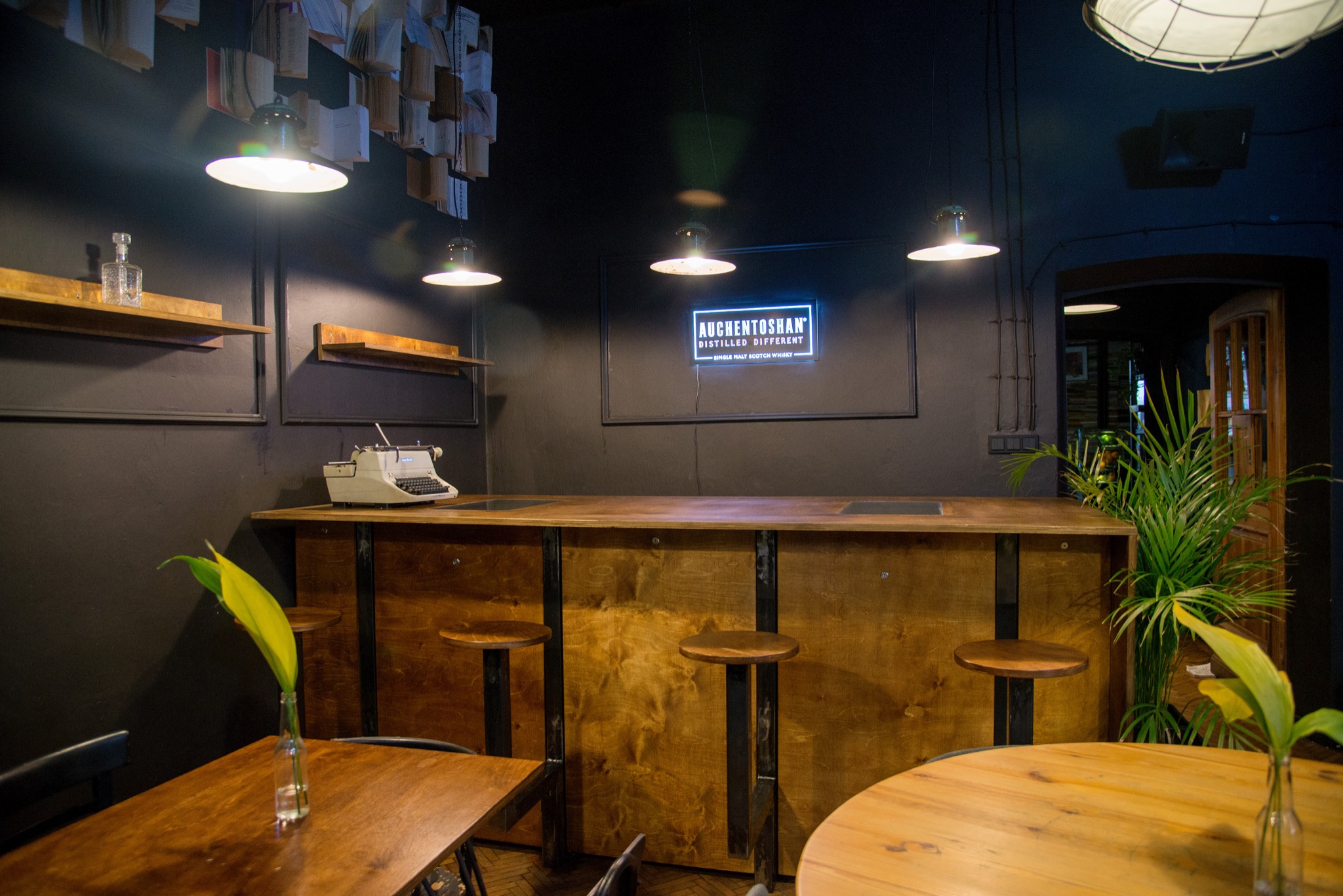 Wnętrze baru w stylu industrialnym z drewnianym blatem, krzesłami barowymi, maszyną do pisania i neonowym szyldem 'Auchentoshan'.