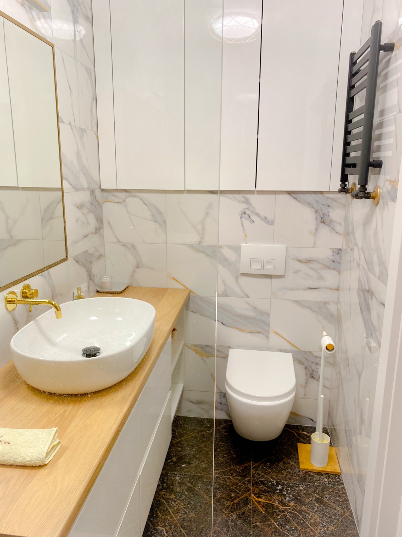 Nowoczesna łazienka z marmurowymi płytkami, owalną umywalką na drewnianym blacie, złotą armaturą i czarnym grzejnikiem drabinkowym.