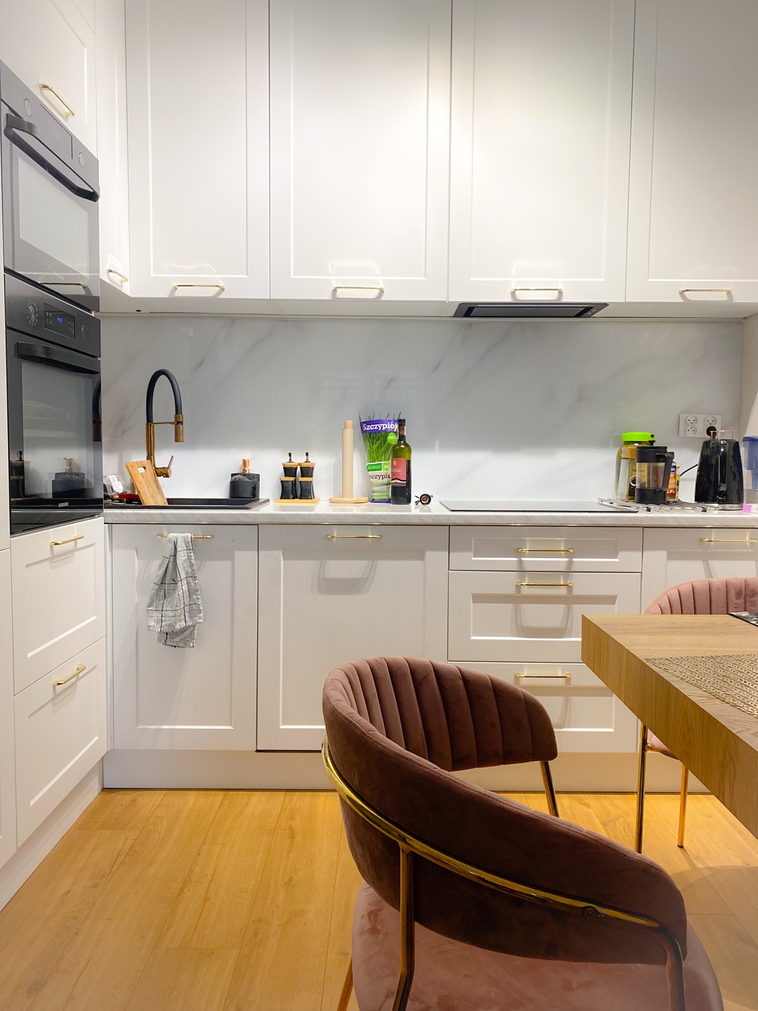 Nowoczesna, jasna kuchnia z białymi szafkami z złotymi uchwytami, marmurowym blatem, czarną armaturą i drewnianym blatem stołu z welurowymi krzesłami w kolorze pudrowego różu.