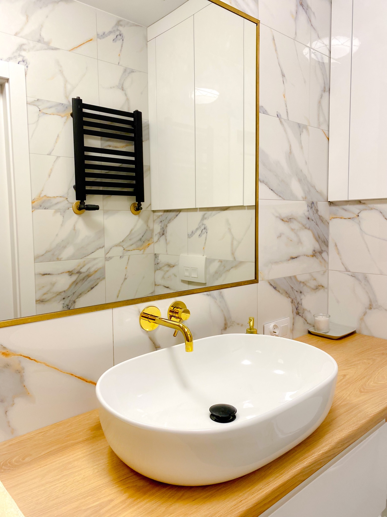Biała, owalna umywalka nablatowa z czarnym korkiem, złota bateria ścienna i marmurowe płytki w łazience z grzejnikiem drabinkowym.