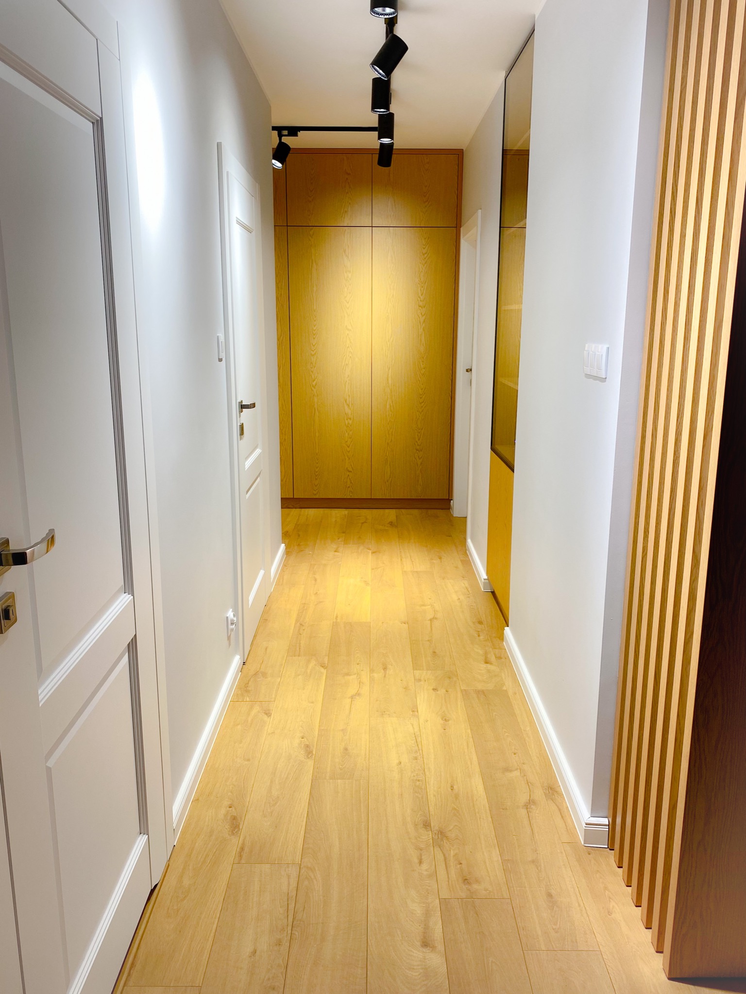Nowoczesny korytarz z drewnianą podłogą, zabudowaną szafą z oświetleniem punktowym, białymi ścianami i dekoracyjnymi lamelami ściennymi.