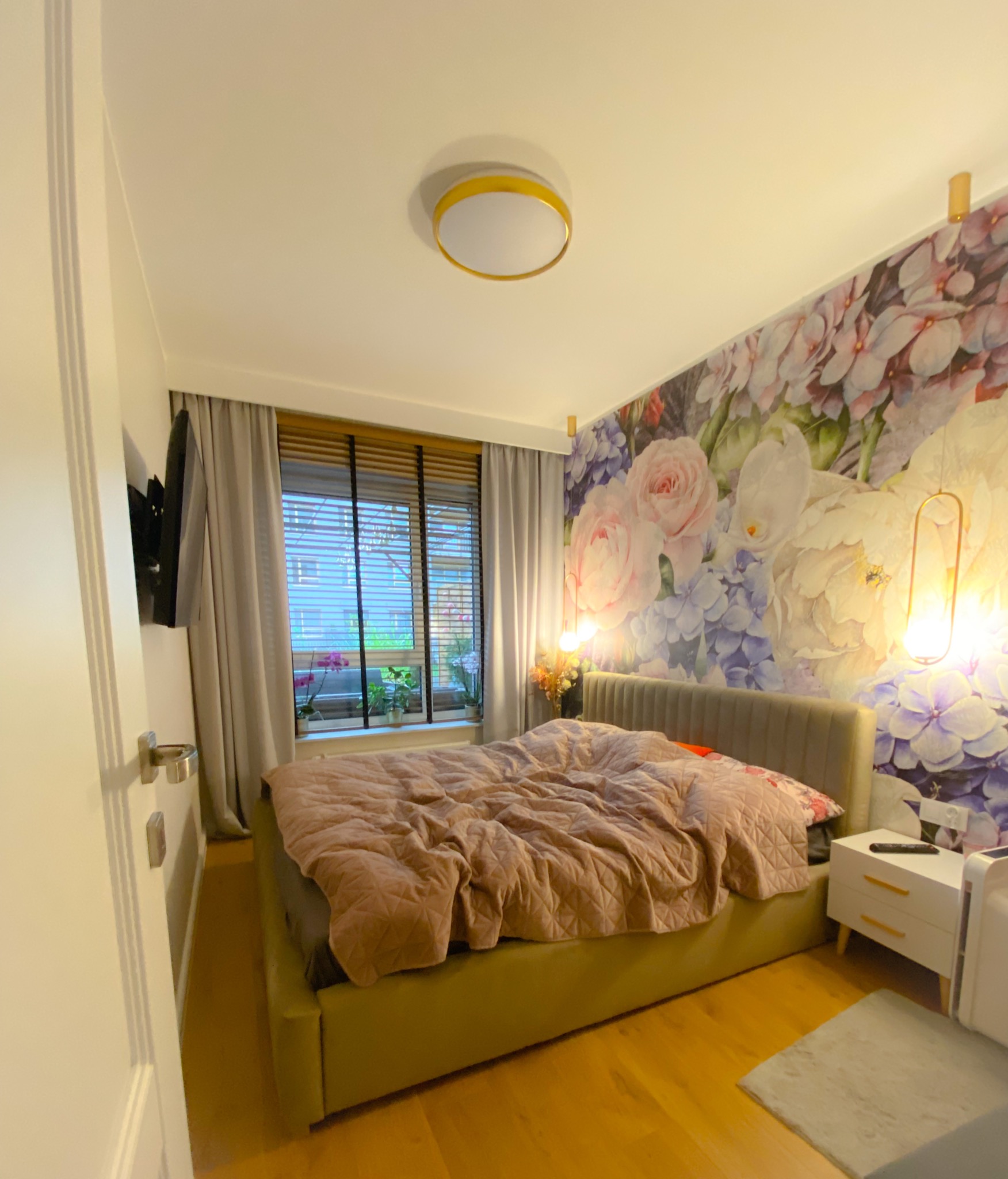 Sypialnia z łóżkiem z zieloną ramą, kwiecista tapeta, lampy wiszące i drewniane żaluzje w oknie.