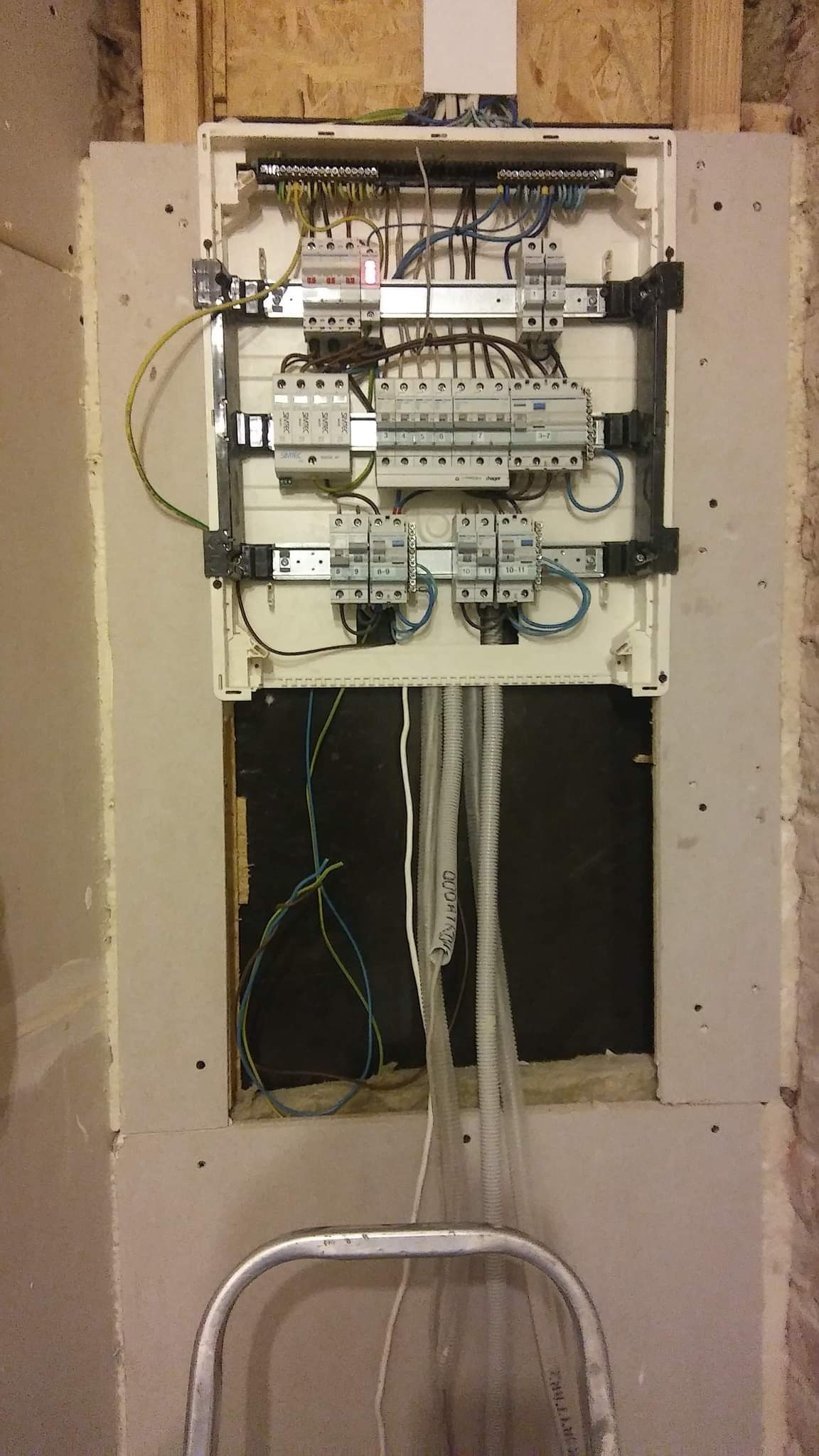 Otwarta skrzynka elektryczna z bezpiecznikami i przewodami w ścianie w trakcie montażu, widoczne rury karbowane i drabina aluminiowa.