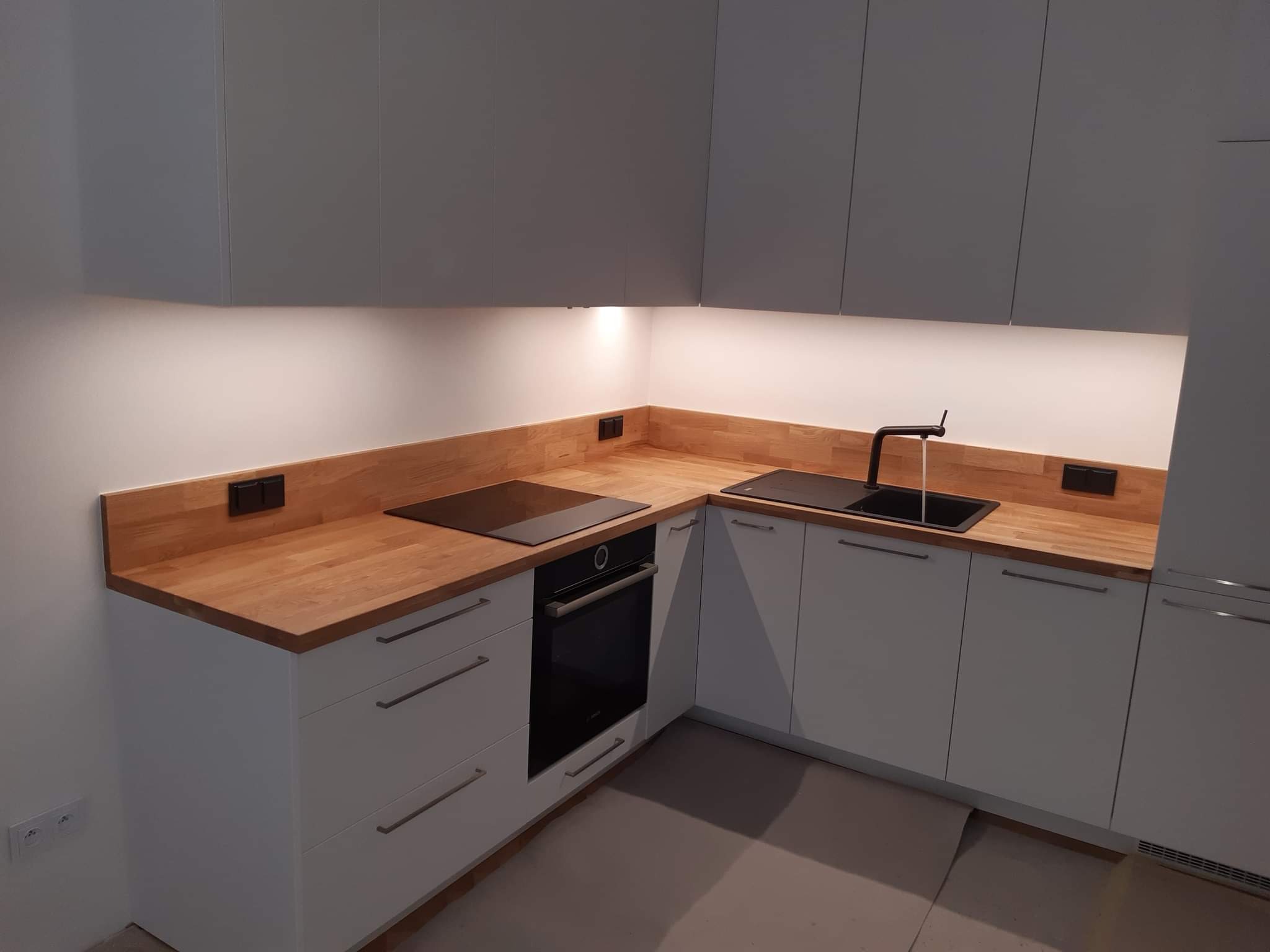 Nowoczesna kuchnia w odcieniach szarości i bieli z drewnianym blatem, czarną armaturą i płytą indukcyjną, oświetlona taśmą LED pod szafkami górnymi.