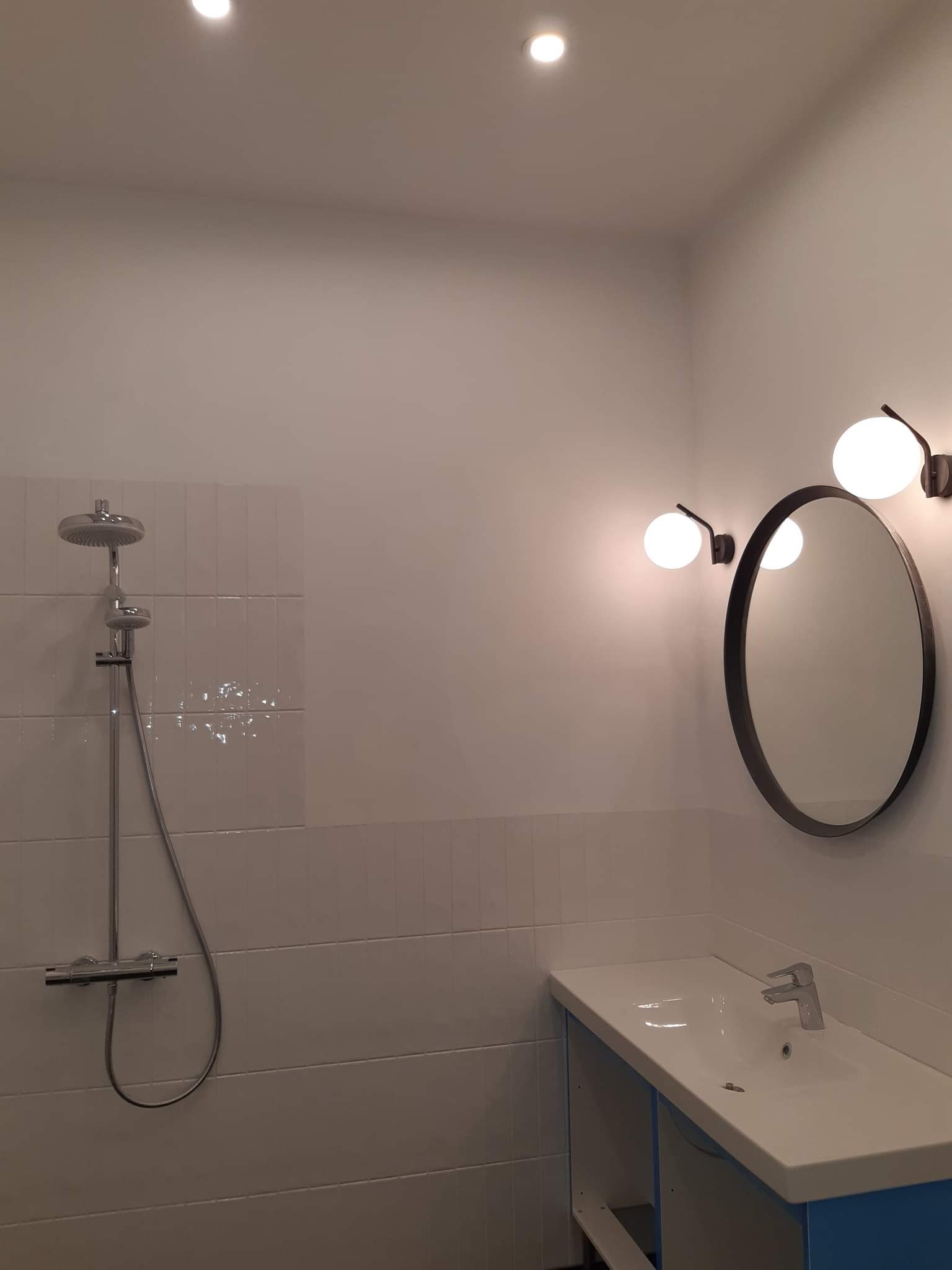 Nowoczesna łazienka z chromowaną baterią prysznicową, okrągłym lustrem z czarną ramą i kulistymi lampami po bokach, umywalką z szafką w kolorze błękitnym i białymi płytkami na ścianach.