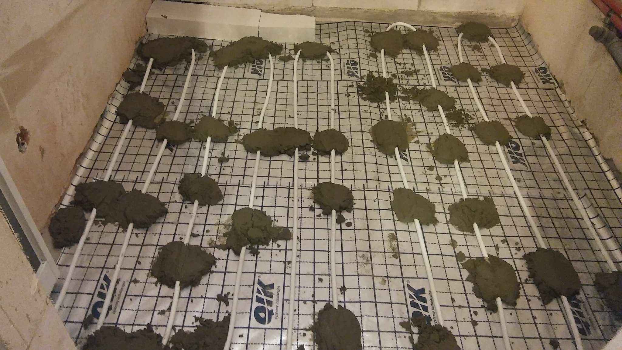 Montaż ogrzewania podłogowego: białe rurki na siatce z nałożonymi punktowo grudkami zaprawy.