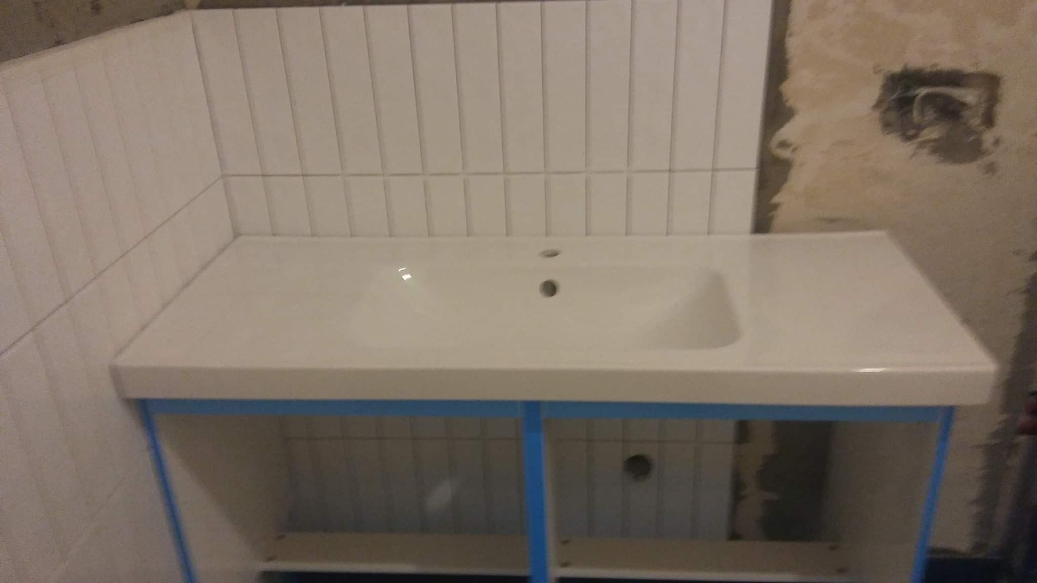 Biała umywalka ceramiczna osadzona na szafce z niebieskimi elementami, zamontowana na tle ściany z białymi, pionowymi płytkami, obok fragment surowej ściany z widocznym otworem na instalację...