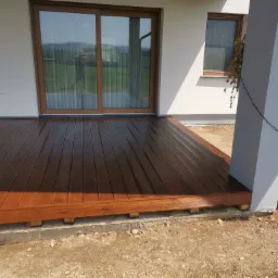 Świeżo polakierowany, brązowy taras drewniany z widocznymi słojami drewna, przylegający do białego budynku z dużym oknem przesuwnym i kolumną.