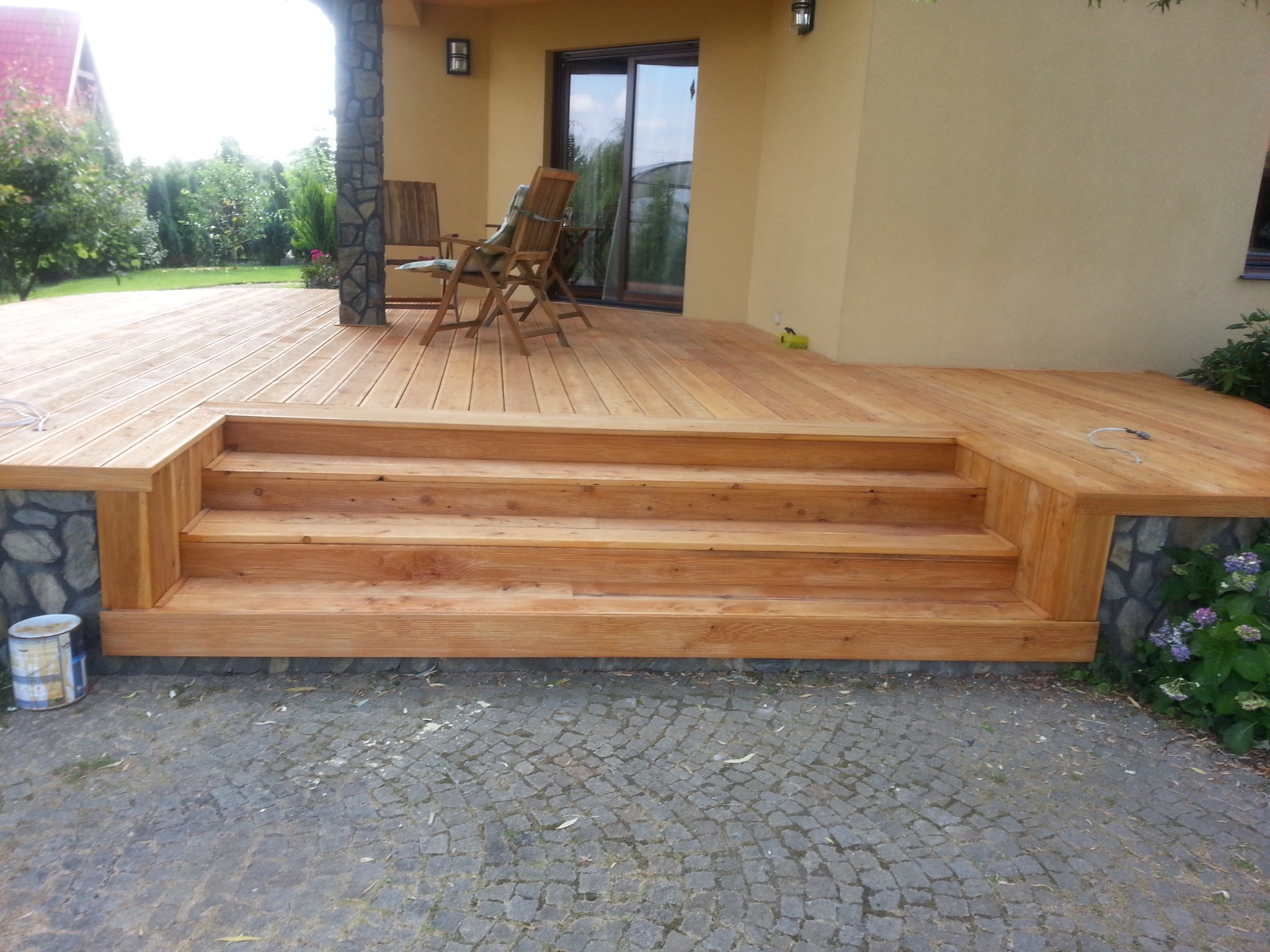 Nowy drewniany taras z trzema schodkami i kamienną obudową, widok z poziomu brukowanej ścieżki.