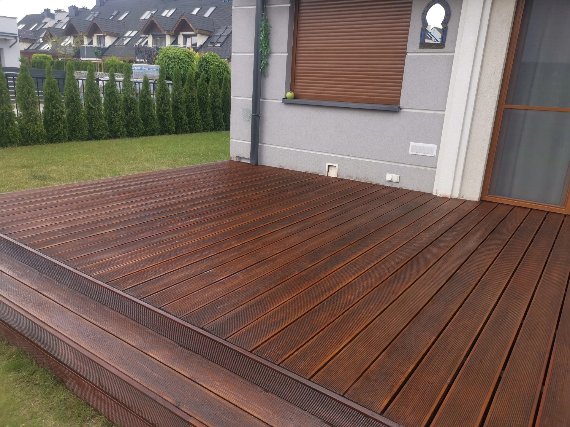 Nowo zaimpregnowany, ciemnobrązowy taras drewniany z ryflowanych desek, przylegający do szarego domu z brązowymi roletami i zielonym jabłkiem na parapecie, widok na trawnik i rząd tuj.