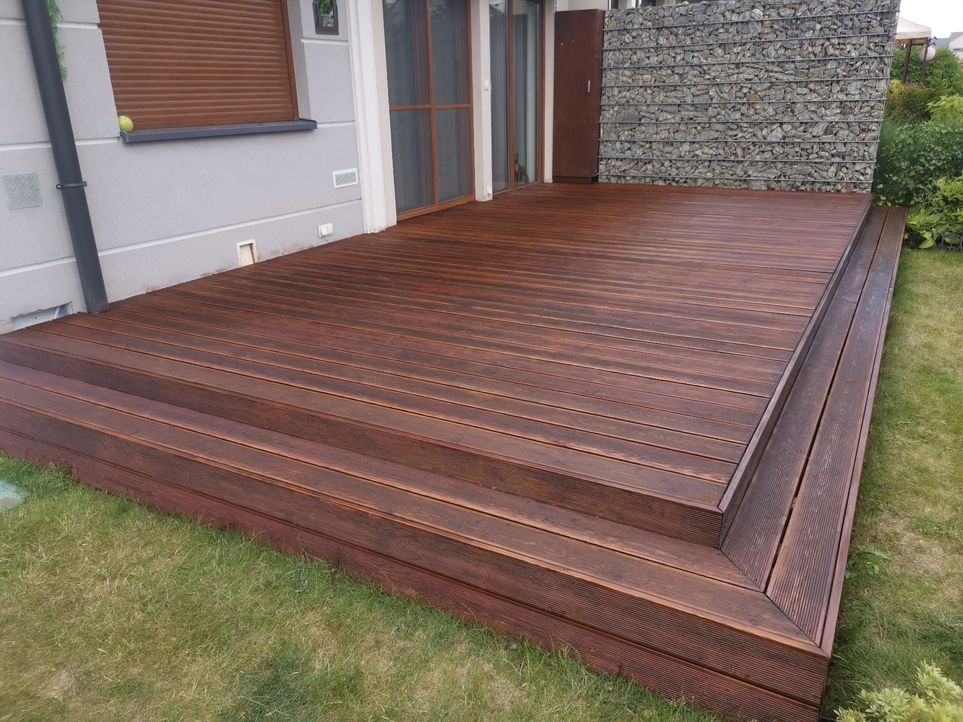 Nowoczesny taras z drewna egzotycznego, wykończony schodkami, z widocznym murem gabionowym w tle.