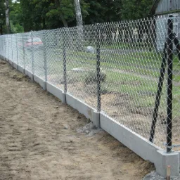 Długi, nowy płot z siatki metalowej na betonowej podmurówce, wsparty czarnymi słupkami, oddzielający posesję od drogi gruntowej, z widocznym samochodem w tle.