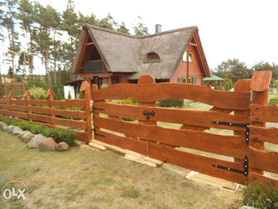 Rustykalny, drewniany płot z bramą, z kutymi zawiasami i zamkiem, na tle domu z trzcinowym dachem i zieleni ogrodu.