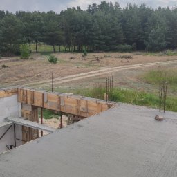 Kdk-bud Kamila Szcześniakiewicz - Wylany betonowy strop na budowie domu jednorodzinnego, widoczne szalunki i wystające pręty zbrojeniowe, w tle las i polna droga.