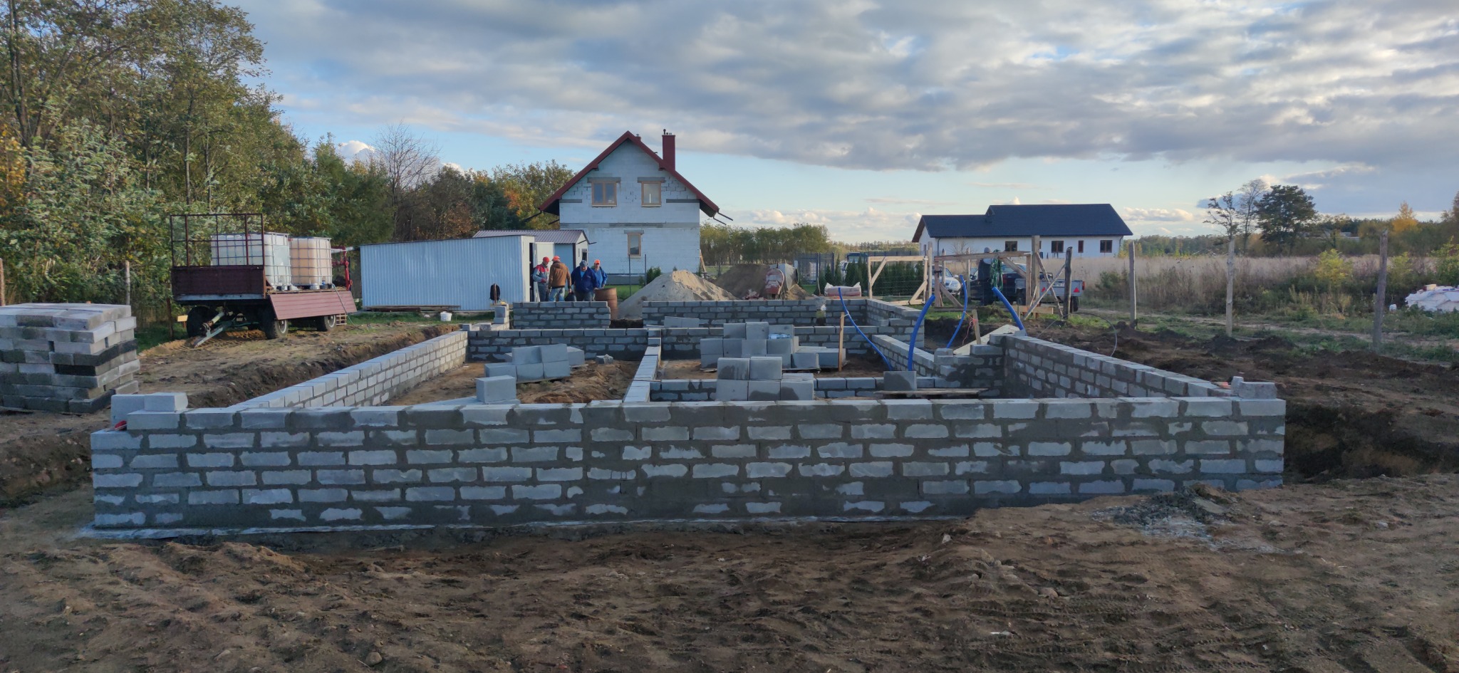 Fundamenty domu z bloczków betonowych, widoczny częściowo wylany fundament, na działce budowlanej, w tle dom w stanie surowym, pracownicy w oddali.