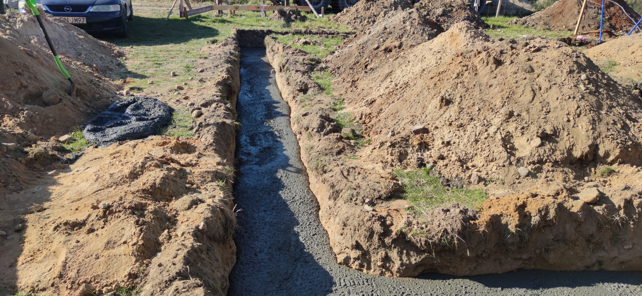Wykopany wąski fundament zalany świeżym betonem, widoczna sterta ziemi i łopata, w tle zaparkowany samochód.
