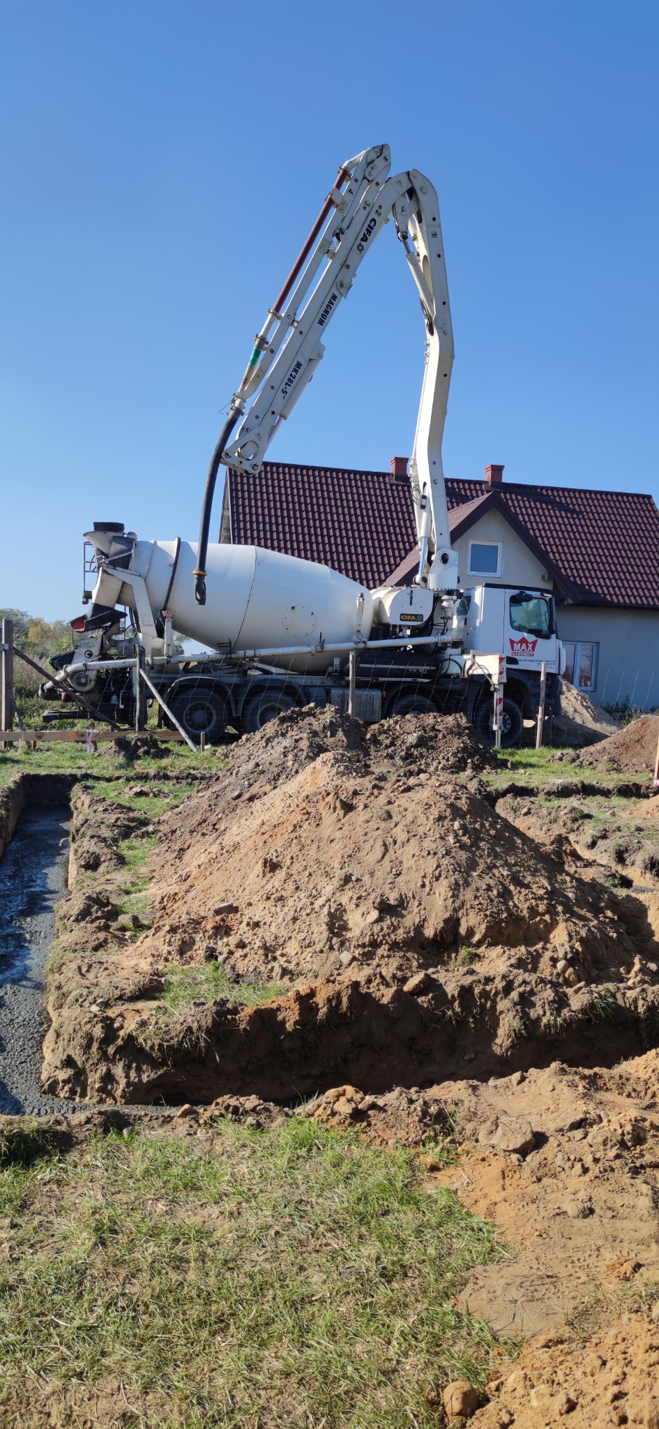 Betoniarka z wysięgnikiem pompuje beton do fundamentów domu jednorodzinnego, widoczna wykopana ziemia i częściowo zalany betonem wykop.