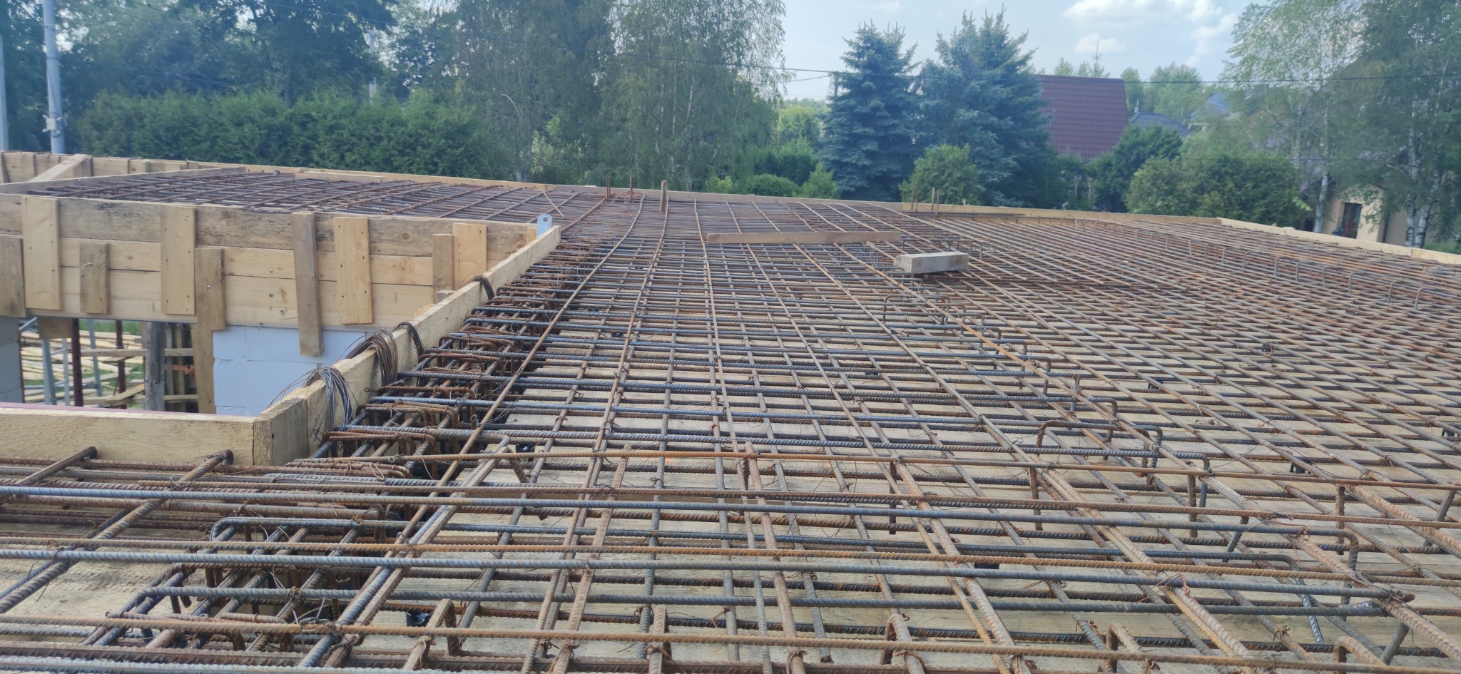 Zbrojenie stropu domu jednorodzinnego, widok z góry na siatkę z prętów stalowych i drewniane szalunki, w tle zieleń drzew i domy.
