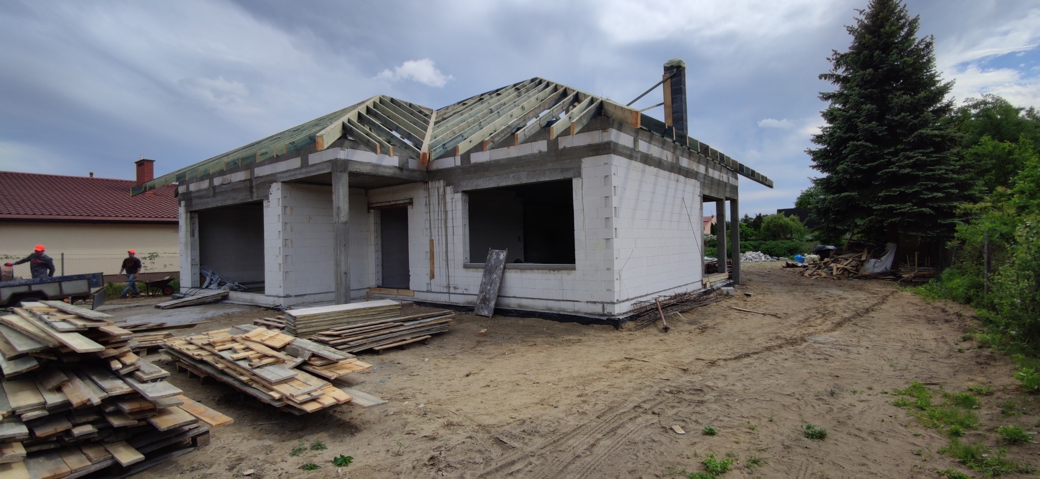 Budowa domu jednorodzinnego w stanie surowym otwartym z widocznym drewnianym szkieletem dachu, białymi ścianami z betonu komórkowego, stertą desek i pracownikami w oddali.