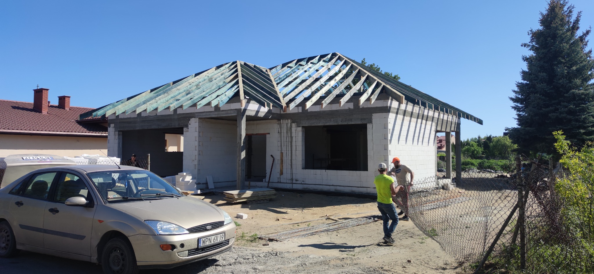 Budowa domu jednorodzinnego w stanie surowym otwartym, widoczne ściany z bloczków, drewniana konstrukcja dachu, dwóch pracowników i zaparkowany samochód osobowy.
