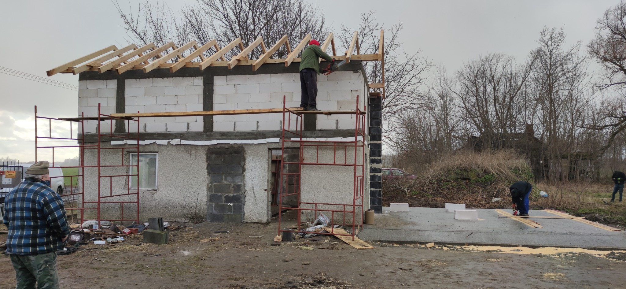 Budowa domu z białych bloczków: murarz na rusztowaniu montuje drewnianą konstrukcję dachu, w tle widoczne drzewa i częściowo zrujnowany budynek.