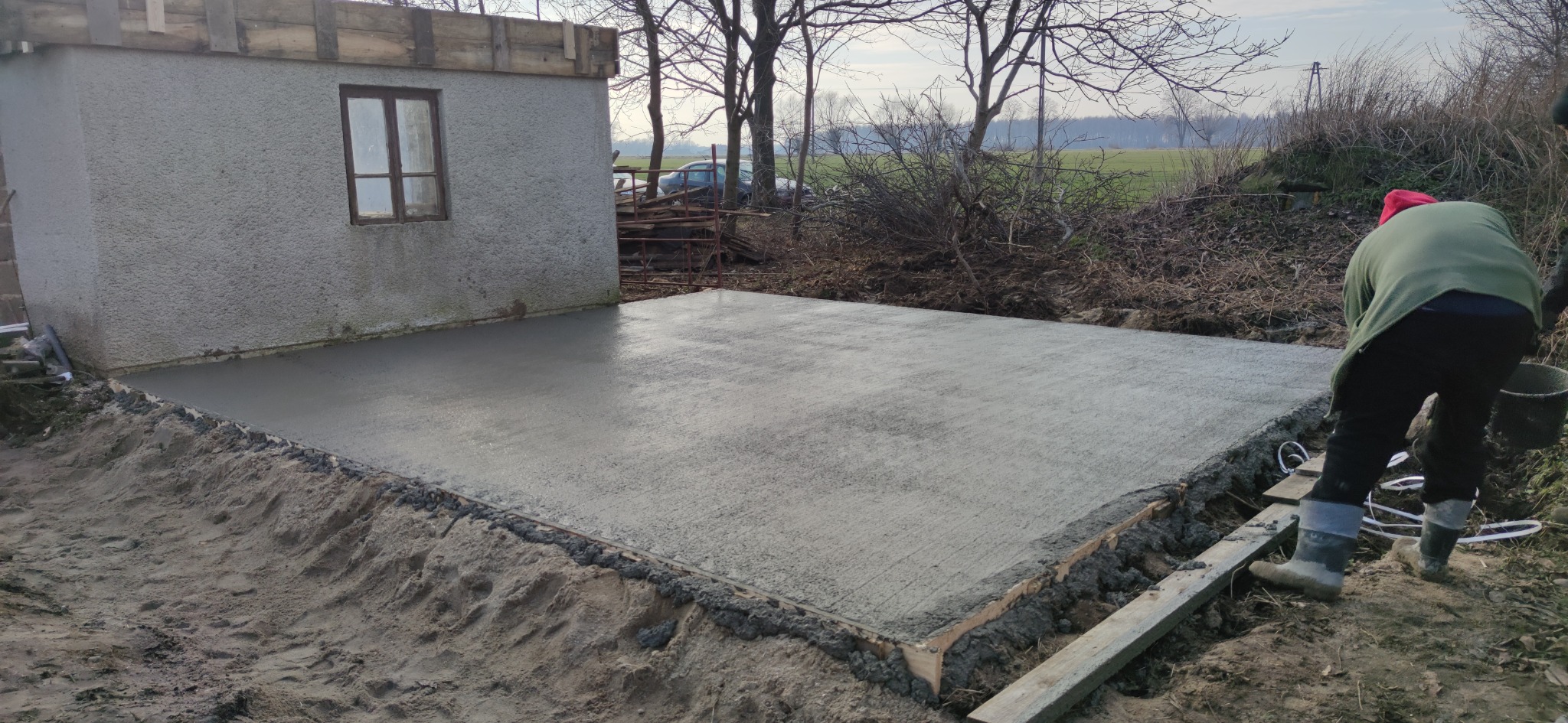 Wylewka betonowa na gruncie, przygotowana pod budowę, widoczny szalunek i fragment ściany budynku z oknem w tle.