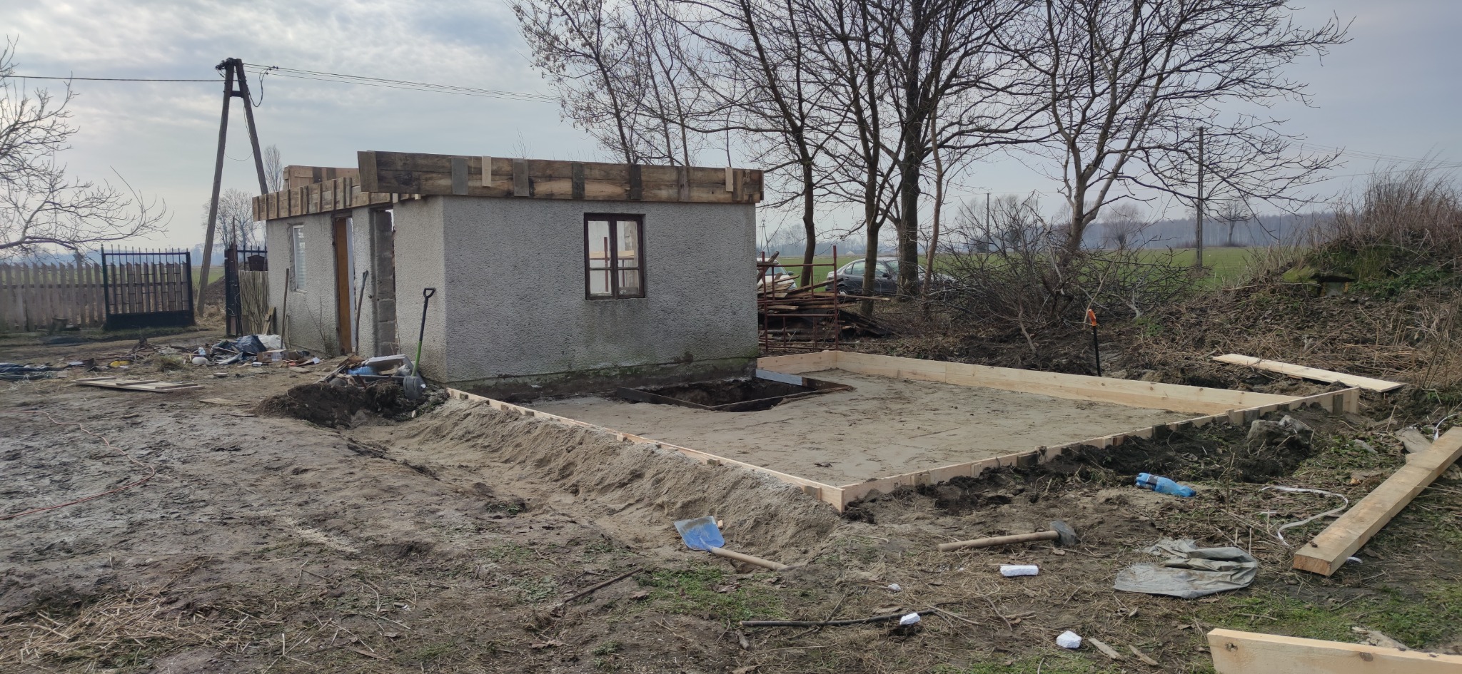 Surowy budynek z szarego tynku z drewnianym szalunkiem na dachu, obok przygotowany fundament pod rozbudowę, widoczna łopata i narzędzia na błotnistym terenie.
