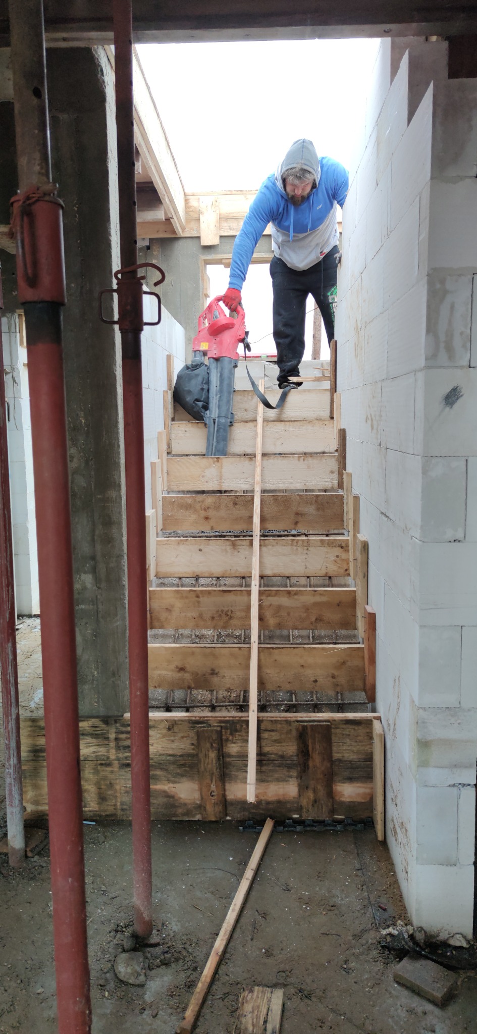 Budowa betonowych schodów wewnątrz domu z bloczków, widoczny pracownik z dmuchawą do czyszczenia szalunku, zbrojenie w deskach, podpory stropu w tle.