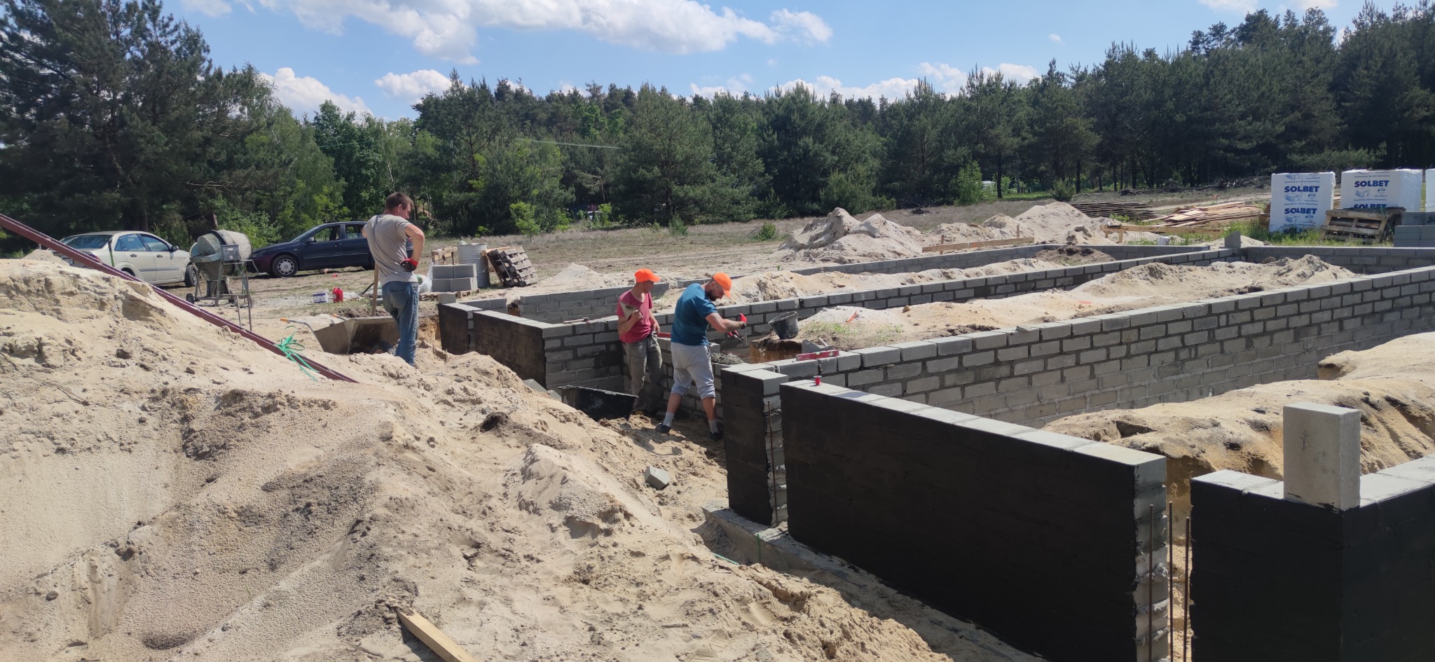 Budowa fundamentów domu z bloczków betonowych w Płońsku, widoczni pracownicy w pomarańczowych czapkach murujący ściany fundamentowe, teren budowy z piaskiem i samochodami w tle.