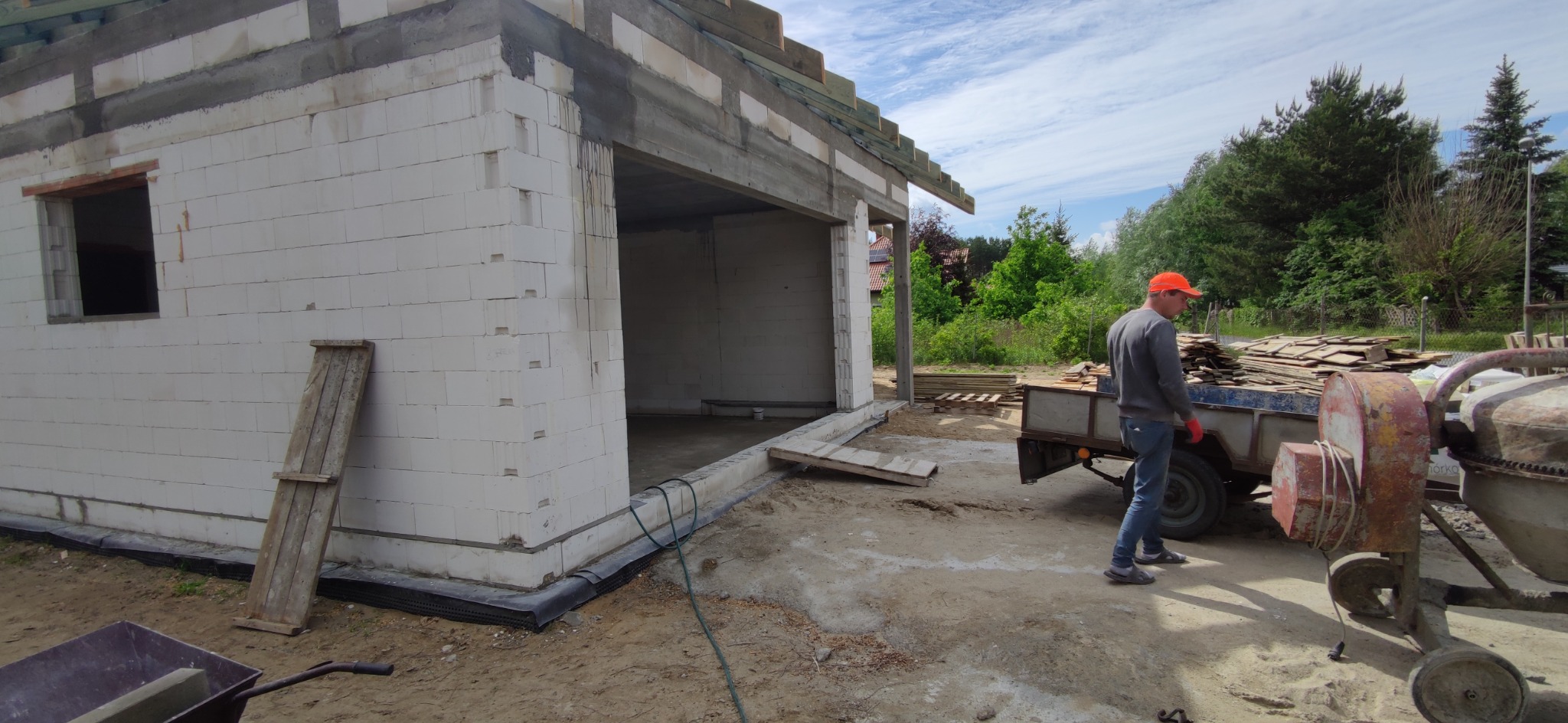 Budowa domu z białych bloczków, widoczny betonowy wieniec, przygotowane krokwie dachowe, przyczepka z dachówką i betoniarka. Pracownik w czerwonej czapce.